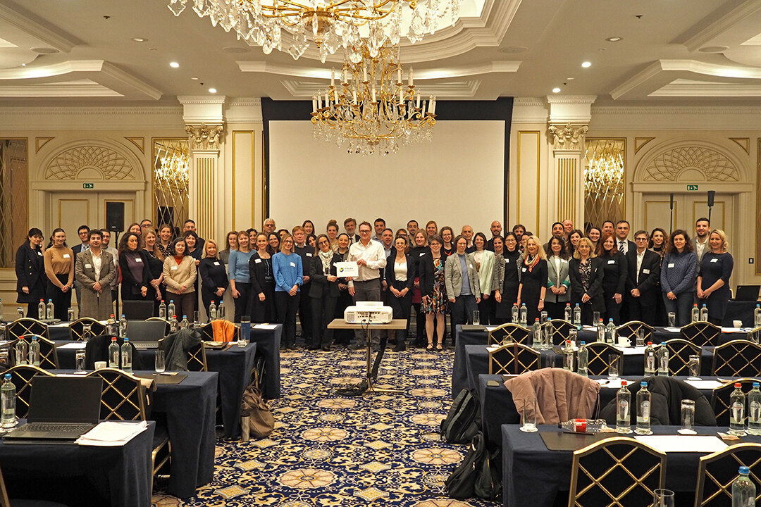 Ein festlicher Saal mit großer Leinwand. Davor stehen ca. 60 Personen, die sich zum Gruppenfoto aufgestellt haben.